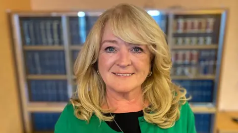 Ms McGahey is standing in front of a book case with leather bound books on the shelves. She has blond wavy hair 