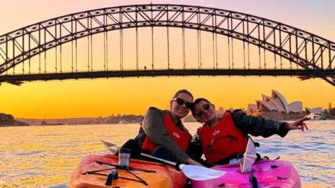 Rosie Scott Fotografia de Rosie Scott (à direita) praticando canoagem no rio Parramatta, em Sydney. Ela está sentada em uma canoa rosa, com um amigo à sua esquerda em uma canoa laranja. Ambos usam óculos escuros pretos e sorriem para a câmera. Rosie estende a mão direita para o lado do corpo e se inclina na direção da amiga. Ambos usam coletes salva-vidas vermelhos. Ao fundo você pode ver a Ópera de Sydney. O sol se põe ao fundo, criando um tom alaranjado no céu.