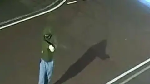 Person in green jacket with hood up and blue jeans standing on a road with a gun in his hand.