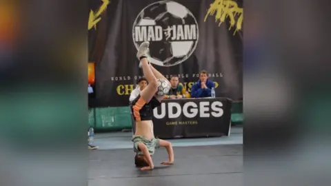 Yvonne Taylor James doing a handstand with a football between his knees. A panel of judges sit on a table behind him, with a black curtain behind them which says Mad Jam inside a football graphic.