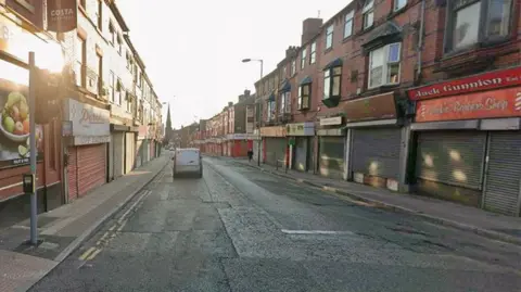 A white van travelling down Breck Road on a clear day. There are a number of shops with shutters down lining the road.