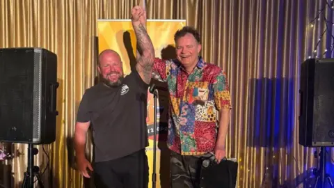 Russ Lee Two men standing on a stage both holding one arm up in the air. The man on the left has a bald head and a beard and is wearing a black t-shirt and black trousers. The man on the right is wearing a colourful patchwork short sleeved shirt. Both men are smiling. 