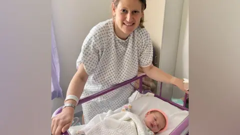 Theo Clarke A woman stands with her hands on the purple frame of a small bed which has a small baby lying in it. She has dark blonde hair, pulled behind her head and wears a hospital gown of white with a dark diamond pattern on it. The baby has a white blanket across it.