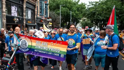 People marching during Notts Pride carrying a rainbow banner which reads "We are Singing Proud"
