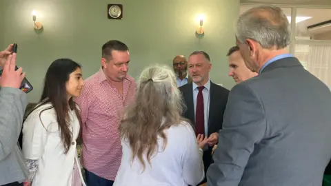 Shariqua Ahmed/ BBC A group of people - including a woman in white, a man in a red checked shirt, another woman who is facing her back to camera and Andrew Pakes - all chatting to Steve Reed.