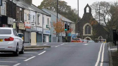 View of Eston High Street leading up to a curve with a church on the top right. A number of closed or empty shops can be seen on the left with a partial view of a white parked car. 