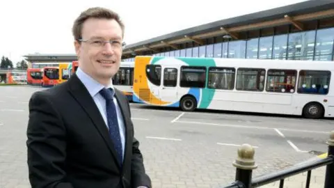 Stagecoach East Midlands Managing Director Matt Cranwell standing to the left of the photo. He is middle-aged wearing a black blazer, light blue shirt and navy tie with white spots. He is smiling, has dark hair and is wearing silver thin framed glasses. He is in a coach park, with a row of yellow, white and turquoise single decker buses behind him. 