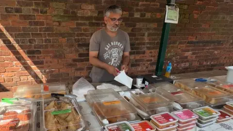 Local Democracy Reporting Service Stallholder Shan Ajaib at Pudsey Market putting food products into paper bags with brightly coloured boxes on his stall. He is wearing a brown t-short with white writing on it and he has a beard