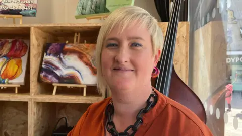 Isabella Verona/BBC Becky Carrier smiling at the camera. She has blonde hair and blue eyeliner and is wearing an orange top. She has a black chain necklace on and is standing in a room with paintings on the shelves behind.