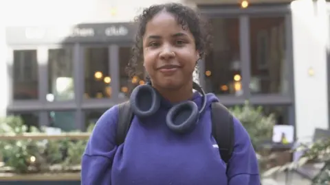 Baya is wearing a purple hoodie and blue headphones around her neck. She has black curly hair which is tied up, with a few loose strands framing her face. She has a nose piercing and a stud below her eye. 