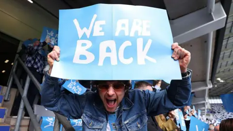 Getty Images Tom Grennan in the stands during the Sky Bet Championship match between Coventry City and Wrexham AFC at The Coventry Building Society Arena on 26 April. He holds up a blue sign with white writing saying We Are Back. He has his mouth open wide in joy and is wearing a denim jacket and shite t-shirt, with dark sunglasses, though his face is in shadow from the sign.