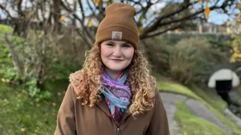 Millie is standing under trees with a stone bridge in the background. She has long curly strawberry blonde hair and is wearing a brown coat, brown hat and multi-coloured scarf. 