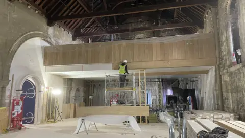 Shaun Whitmore/BBC A workman stands on a scaffolding platform as he continues work inside Norwich Castle's keep. He is working on what appears to be a section of wooden panelling. The vaulted ceiling of the keep is above him. 