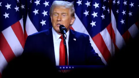 Donald Trump addresses supporters at an election night watch party in the West Palm Beach Convention Center in West Palm Beach, Florida, on 6 November. He is standing at a podium with the stars and stripes of the American flag behind him. Trump is wearing a blue suit, white shirt and red tie and has a microphone near his mouth