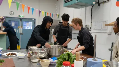 Three young people in black aprons are preparing food on a kitchen island. Pots and pans are on the island with ingredients.  