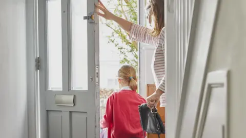 A child in a red school uniform walking out a door. A woman holding a black back is holding the door open.