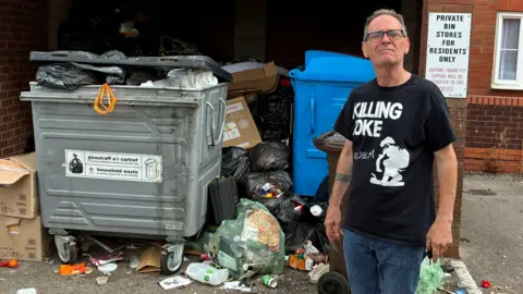 David Power in front of overflowing containers with waste dumped in bags and littered across the tarmac next to wheelie bins in bin store area. He wears a black t-shirt and shorts and has short grey hair and wears glasses.