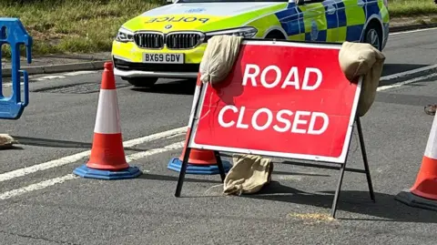 A road closure sign in front of a police car. There are three orange cones around the red sign. 