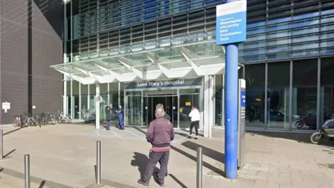 Google A sign above the entrance to the hospital says 'Saint Mary's Hospital', with several people walking or standing near to it. A large paved area is in front of the entrance doors.