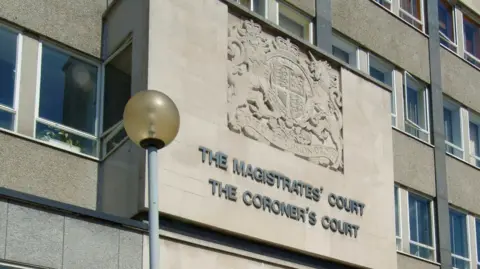 Exterior view of the stone facade of a building featuring the carved text "The Magistrates' Court" and "The Coroner's Court," with a royal coat of arms above and a streetlamp in the foreground.
