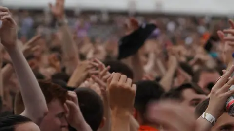 BBC A generic photo of a crowd at an outdoor festival. People's hands are raised in the air.