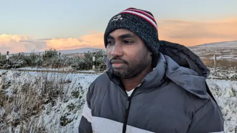 a man in a beanie hat and thick jacket looking away from the camera, with snowy fields around him and a hill in the background