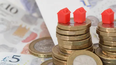 A pile of coins with red monopoly board game houses sat on top. In the background there are £5, £10 and £20 notes.