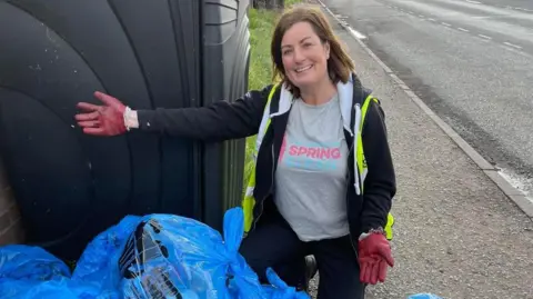 Karen Blanchfield Karen Blanchfield has shoulder-length brown hair and is smiling. She is wearing a grey T-shirt and black hoodie and yellow high-visibility vest. She is kneeling next to a pile of blue plastic bags and a large black plastic bin.