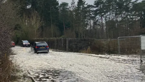 Flooding in Taverham. Water has settled on the road and cars are passing through the water.