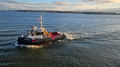 A harbour tug travels across the water outside of the Port of Workington. The tug has HELVELLYN written on the bow of the black boat and it has a bright red floor and white cabin. 