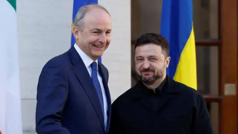 Reuters Micheál Martin stands beside Zelensky outside Government Buildings in Dublin. The two men are smiling as they pose in front of Irish tricolours and the Ukrainian flag. Martin is wearing a navy suit and tie with a white collared shirt. Zelensky is wearing a black coat with a black shirt underneath.