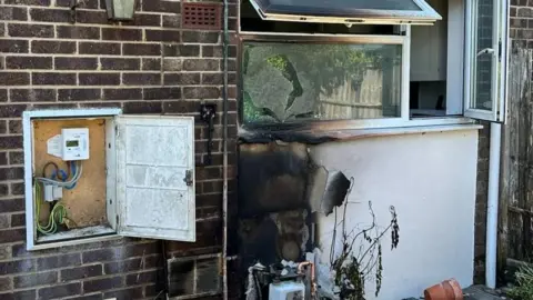A burnt section of a wall beneath a window. There is a gas meter nearby and another meter on the ground beneath the burnt area