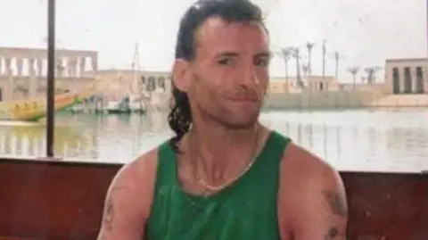 south wales police Timmy sat on a boat looking at the camera with a slight grin. He is wearing a green vest top and a chain necklace. He has a dark brown mullet and has a few tattoos on his arms. 