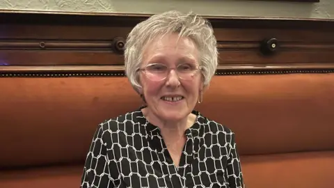 Older woman, Linda with grey hair and glasses sits on a brown leather bench seat and smiles. She wears a black and white blouse.
