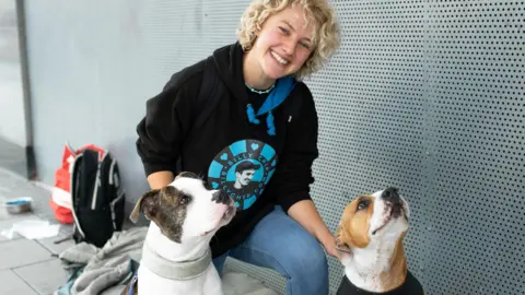 A woman is smiling as she kneels down to take a photo with two bulldogs. She wears a t-shirt with the Billy Chip logo and blue jeans. Her hair is short and blonde. She seems happy. They are outside and in the background there is a water bowl, most likely for the two dogs who are most likely owned by a homeless person.