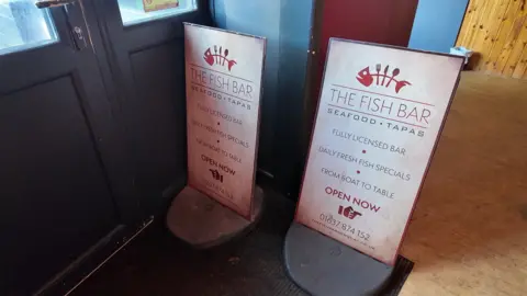 A close up image of two street-side restaurant signs that are inside the front door of a restaurant.  They say The Fish Bat, Seafood, Tapas.   