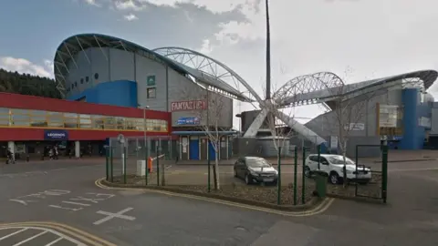 The outside of Huddersfield's John Smith's Stadium showing the back two of its arched stands, with an access road and car parking area in front.