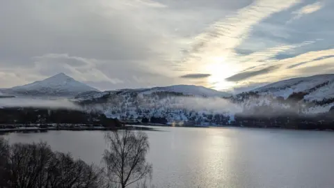 Angela Jones A view of Schiehallion from the window of a hotel room. The mountains are covered in snow. There are trees on the landscape which look dark. The sun is coming up.