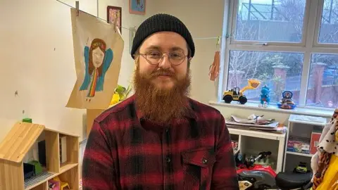 Aaron pictured with a black beanie hat on and has a long ginger beard. He is wearing a black and red plaid shirt and thin gold rimmed circular framed glasses. 