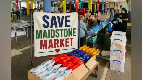A sign saying 'Save Maidstone Market' at Maidstone Market. The sign is in blue, green and red and a number of stalls and visitors can be seen behind it.