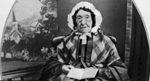 National Musuems Northern Ireland A black and white photo of an elderly lady sat on a chair. She is wearing a white bonnet on her head, a chequered scarf and long frock. She is holding an open book and looking directly at the camera.