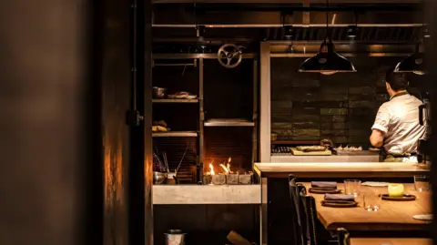 Eleonora Boscarelli Chef Corrin with seen from behind in white uniform preparing food in open kitchen. Tables laid for guests are visible just on the other side and to his left are shelves and a small open fire for cooking.