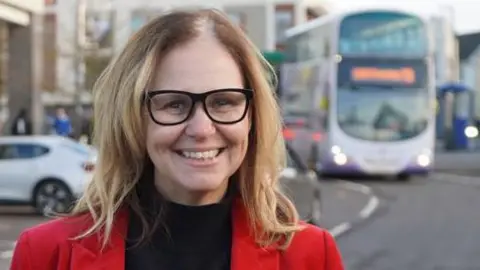 Weca Helen Godwin standing by a road in a red jacket, black top and black glasses. In the background there is a bus to her right, driving along the road.