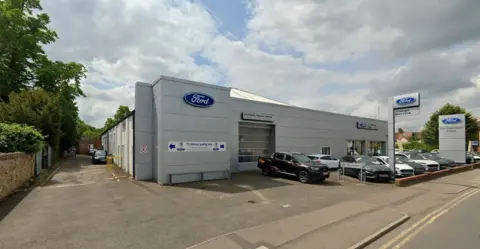 Google A Ford car garage. There are cars on the forecourt and large blue Ford signs on the grey building.