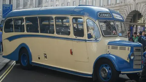 Robert Lamb/Geograph Vintage bus exhibition. People are looking at a blue and cream coloured bus. It has two large headlights at the front and a bonnet with air grills. A metal fuel filler cap can be seen on the vehicle's right side.
