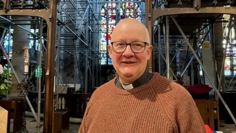 Rev Holliday, who wears glasses and brown jumper over his clerical collar, smiles as he stands in front of scaffold with a large stained glass window behind.