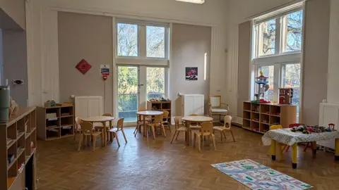 Wescott Tots The new room at Holderness House. There are large windows, a brown parquet floor with shelves at the side and desks and chairs in the middle