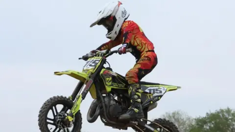 supplied A young motorbike rider jumps in the air on his bike. He wears an orange jumpsuit and white helmet.