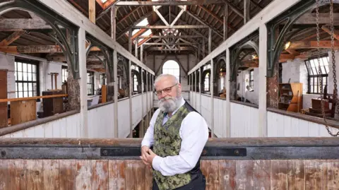 Historic England Inside an old building with a wooden interior a man stands with his elbow leaning against a side of a balacony. He is smiling at the camera and wearing a green waistcoat. 