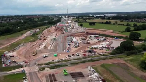 A brown scar of land surrounded by green fields shows a construction site. The picture is shot from above, various roads and construction vehicles can be seen.